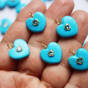 May include: A collection of bright turquoise heart-shaped charms, each featuring a small, centered heart-shaped gemstone set in a gold-toned metal bezel. Each charm includes a small gold-toned jump ring attached to the top for hanging purposes.
