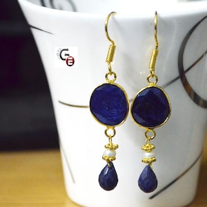 以下が含まれることがあります： 青い宝石の雫がついた、金のぶら下がりイヤリングのペア。イヤリングは、ファセットデザインの丸い青い宝石と、その下にぶら下がっている小さな涙型の青い宝石が特徴です。