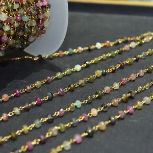 May include: A gold chain with small, faceted, multi-colored gemstones. The chain is made of gold-plated metal and is strung with small, faceted gemstones in various shades of pink, green, and black. The chain is laid out in a straight line on a dark surface.