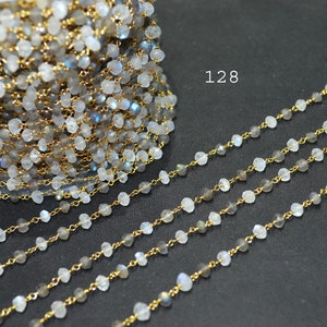 May include: A strand of gold chain with white moonstone beads. The chain is shown in a close-up view, with several strands laid out on a dark surface. The number 128 is visible in the image.