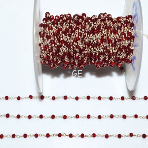 Puede incluir: Un carrete de cadena de cuentas de vidrio rojo con eslabones de cadena plateados. La cadena está dispuesta en tres filas sobre una superficie blanca.