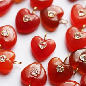 May include: A collection of heart-shaped pendants made of smooth, translucent red stone. Each pendant features a small, clear, triangular-cut gemstone set in a gold-toned metal bezel at the center, with a small gold-toned jump ring attached at the top.