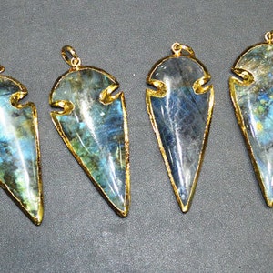 May include: Seven labradorite pendants with gold-colored metal trim. The pendants are shaped like arrowheads, with a polished, iridescent surface displaying blue, green, and yellow hues. Each pendant has a small loop at the top for a chain.