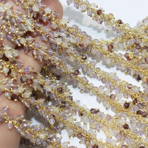 May include: A delicate gold-toned chain adorned with numerous small, faceted, translucent beads in shades of pale purple, light yellow, and deep brown. The beads are densely clustered along the length of the metallic chain, creating a textured, shimmering appearance throughout the entire piece.