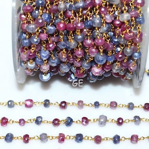 Op de afbeelding: Een spoel met een gouden ketting met afwisselend blauwe en roze facetgeslepen edelsteen kralen. De ketting ligt in drie rijen op een wit oppervlak.