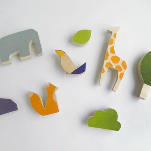 Puede incluir: Un conjunto de juguetes de madera con temática de animales y naturaleza. El conjunto incluye un elefante gris, una jirafa amarilla y naranja, un tucán azul y amarillo, un zorro naranja, una cueva morada y dos árboles verdes.