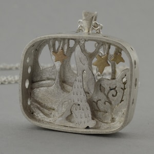 Puede incluir: Un collar de plata con un colgante cuadrado que presenta un diseño recortado detallado de un conejo en un entorno de bosque con detalles dorados. El colgante está unido a una cadena de plata.