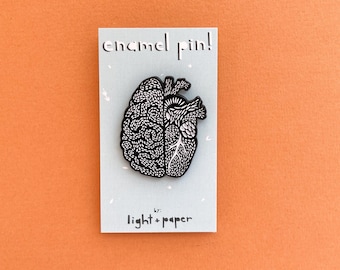 May include: Black and white enamel pin featuring a stylized brain and heart design. The pin reads "enamel pin! by: light + paper".
