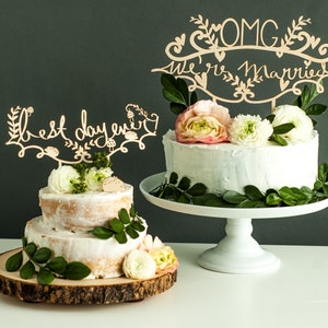 May include: Two wedding cakes decorated with greenery and flowers. The larger cake has a white frosting and a wooden topper that says "OMG We're Married". The smaller cake has a wooden topper that says "Best Day Ever".