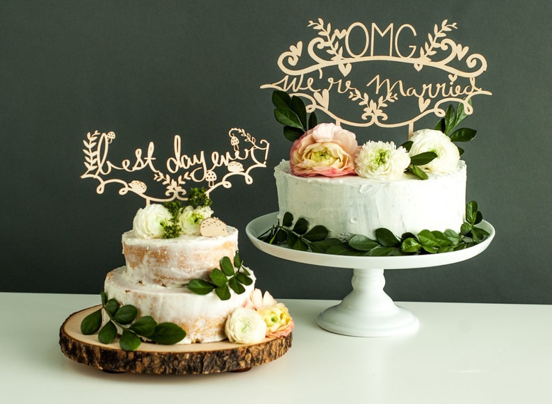 May include: Two wedding cakes decorated with greenery and flowers. The larger cake has a white frosting and a wooden topper that says "OMG We're Married". The smaller cake has a wooden topper that says "Best Day Ever".