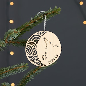 Könnte beinhalten: Ein hölzerner Weihnachtsbaumschmuck in Form eines Halbmondes mit dem Sternbild Fische und dem Wort "Fische" darauf.