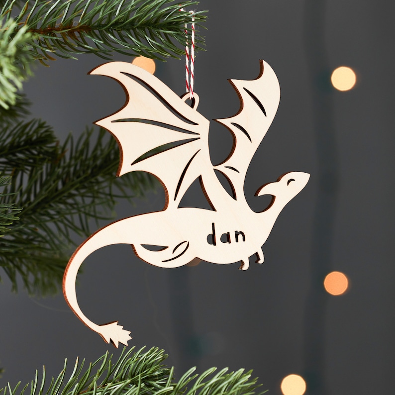 May include: A wooden dragon ornament with a red and white string for hanging. The dragon is flying with its wings spread out and its tail curled. The name "Dan" is written on the dragon's body.