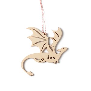 May include: Wooden dragon ornament with a red and white string for hanging. The dragon has wings and a tail and the name "dan" is carved into the body.