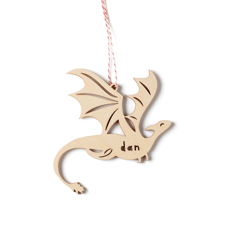 May include: Wooden dragon ornament with a red and white string for hanging. The dragon has wings and a tail and the name "dan" is carved into the body.