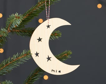 May include: A wooden crescent moon ornament with stars cut out and the name "Mia" written on it. The ornament is hanging from a red and white string on a green Christmas tree.