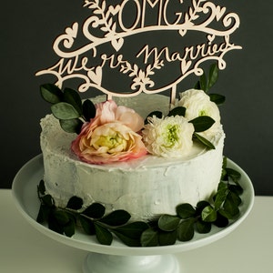 May include: A white wedding cake with light blue frosting decorated with green leaves and pink and white flowers. A wooden cake topper reads "OMG We're Married".