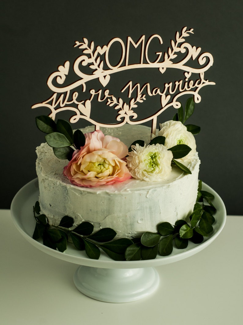 May include: A white wedding cake with light blue frosting decorated with green leaves and pink and white flowers. A wooden cake topper reads "OMG We're Married".
