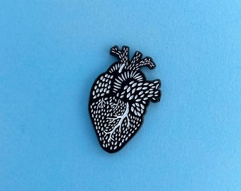 May include: A black and white anatomical heart pin. The pin features a detailed illustration of a human heart, with white veins and textures against a black background. The pin is set against a light blue backdrop.