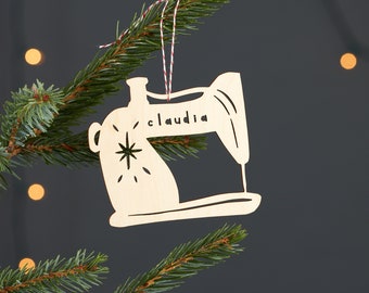 May include: A wooden sewing machine bauble with the name "Claudia" engraved on it. The bauble is hanging from a red and white string on a green Christmas tree.