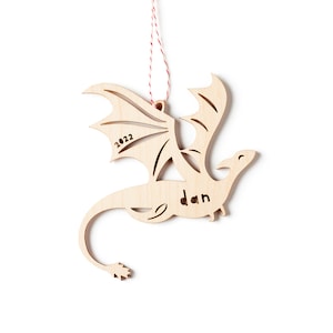 May include: Wooden dragon ornament with a red and white string for hanging. The dragon is flying with its wings spread. The ornament is personalized with the name "Dan" and the year "2022".