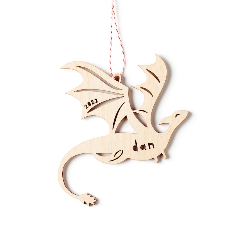 May include: Wooden dragon ornament with a red and white string for hanging. The dragon is flying with its wings spread. The ornament is personalized with the name "Dan" and the year "2022".