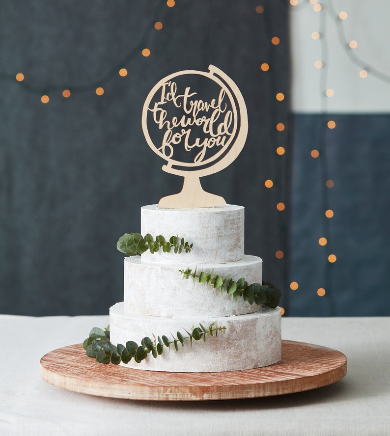 K&ouml;nnte beinhalten: Eine dreist&ouml;ckige Hochzeitstorte, verziert mit Eukalyptusgr&uuml;n und einem Holz-Globus-Tortenaufsatz mit der Aufschrift "I'd travel the world for you".