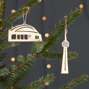 May include: Two wooden Christmas ornaments, one shaped like the CN Tower and the other shaped like the Rogers Centre, both hanging on a green Christmas tree.