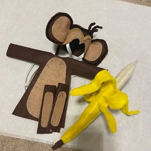 May include: A brown felt monkey costume with a felt banana prop. The costume includes a hat, a body, and two arm pieces. The banana is yellow and has a brown stem.