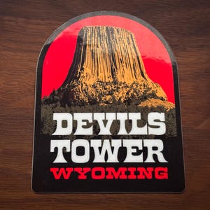 May include: A black and white sticker with a red background. The sticker features a picture of Devils Tower National Monument in Wyoming. The text "Devils Tower Wyoming" is printed in white letters.