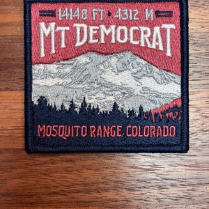 May include: Embroidered patch featuring a mountain scene with the text "MT DEMOCRAT" in red and white. The patch also includes the text "MOSQUITO RANGE, COLORADO" and the elevation "14148 FT • 4312 M". The patch has a navy blue border.