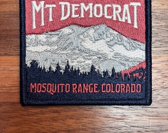 May include: Embroidered patch featuring a mountain scene with the text "MT DEMOCRAT" in red and white. The patch also includes the text "MOSQUITO RANGE, COLORADO" and the elevation "14148 FT • 4312 M". The patch has a navy blue border.