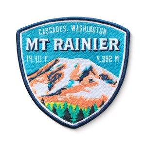 May include: A blue and white embroidered patch with the text "CASCADES WASHINGTON MT RAINIER 14,410 FT 4,392 M" and a depiction of a snow-capped mountain.