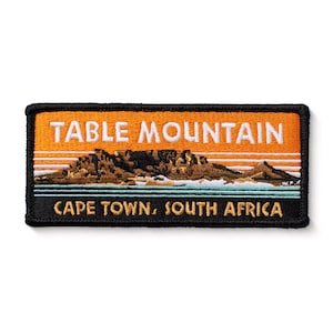 May include: An orange and black embroidered patch with the text "TABLE MOUNTAIN" in white and "CAPE TOWN, SOUTH AFRICA" in gold. The patch features a brown and green embroidered image of Table Mountain.