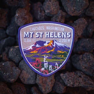 Puede incluir: Un parche bordado morado y azul con el texto "Cascades, Washington, Mt St Helens, 9,366 ft, 2,550 m" y una ilustración colorida de una montaña.
