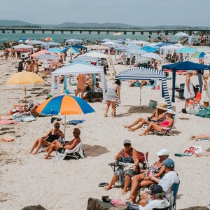 May include: A crowded beach with people relaxing under colorful striped umbrellas. Some people are sitting in chairs, while others are swimming in the ocean. There is a bridge in the background.