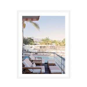 May include: A view from a balcony overlooking a swimming pool with lounge chairs and a palm tree in the foreground. The pool is surrounded by white buildings and lush greenery.