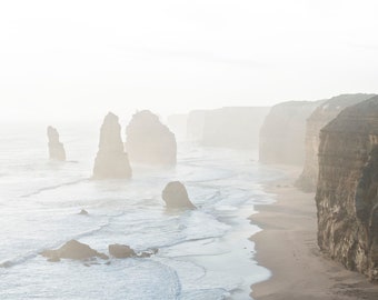 May include: A misty, foggy landscape with tall rock formations rising from the ocean. The water is a light blue and the sky is a pale white. The rocks are a dark brown and are weathered and eroded.
