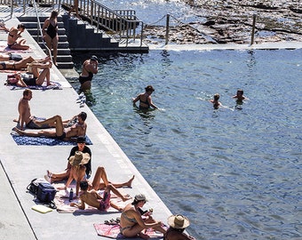 May include: A group of people relax and swim in a pool of clear blue water. Some people are sunbathing on the edge of the pool, while others are swimming in the water. The pool is surrounded by a concrete deck and a rocky shore.
