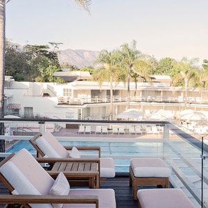 May include: A view from a balcony overlooking a swimming pool with lounge chairs and a mountain range in the background. There are three wooden chairs with pink cushions and a glass railing.