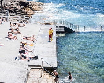 May include: A view of a public outdoor pool with people swimming and sunbathing. The pool is located on a rocky cliff overlooking a beach and the ocean. The water is clear and blue.