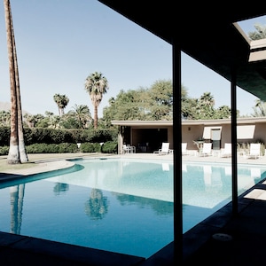 May include: A swimming pool with a curved edge, surrounded by palm trees and a modern building with a covered walkway. The pool is surrounded by a lush green lawn and a hedge.