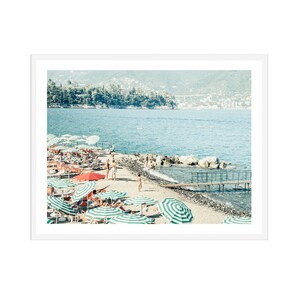 May include: A beach scene with green and white striped umbrellas and red umbrellas, with people relaxing on lounge chairs. The beach is sandy with a wooden walkway leading to the water. The water is blue and there are hills in the background.