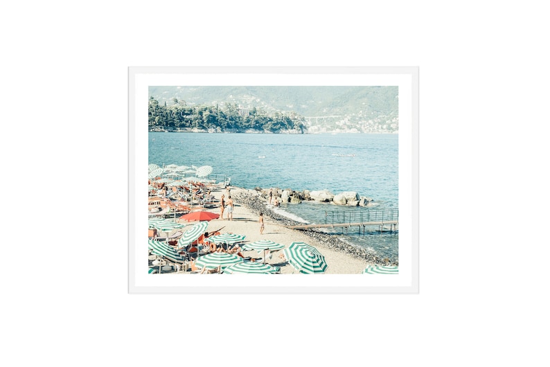 May include: A beach scene with green and white striped umbrellas and red umbrellas, with people relaxing on lounge chairs. The beach is sandy with a wooden walkway leading to the water. The water is blue and there are hills in the background.