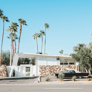 May include: A black vintage car parked in front of a white building with a stone wall. The building has a large overhang and the number 2301 is visible on the side. Palm trees line the front of the building.
