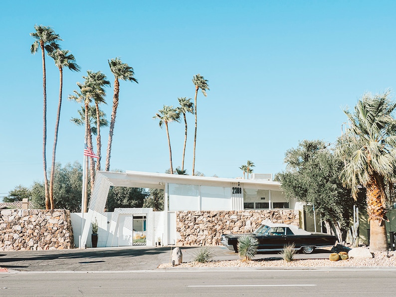 May include: A black vintage car parked in front of a white building with a stone wall. The building has a large overhang and the number 2301 is visible on the side. Palm trees line the front of the building.