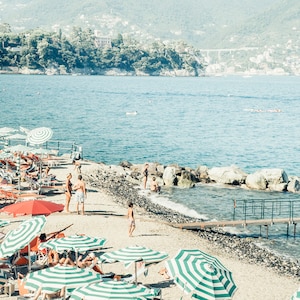 May include: A beach scene with striped umbrellas and lounge chairs on a pebble beach. People are swimming in the clear blue water. There are rocks and a wooden walkway leading to the water.