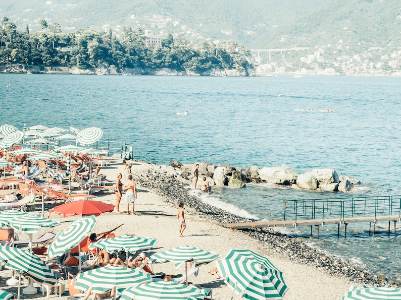 May include: A beach scene with striped umbrellas and lounge chairs on a pebble beach. People are swimming in the clear blue water. There are rocks and a wooden walkway leading to the water.