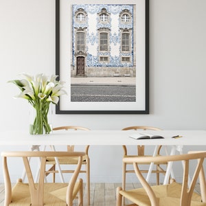 May include: A framed photograph of a building with blue and white tile work. The building has three windows and a door. The photograph is hanging on a white wall above a white table with four wooden chairs.