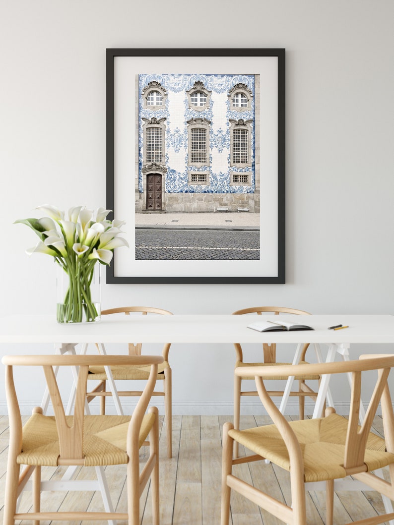 May include: A framed photograph of a building with blue and white tile work. The building has three windows and a door. The photograph is hanging on a white wall above a white table with four wooden chairs.