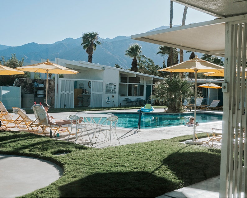 May include: A swimming pool with yellow umbrellas and white sun loungers. The pool is surrounded by green grass and palm trees. There are mountains in the background.